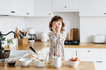 Cute 5 years girl wearing dress preparing cake at the white modern kitchen.