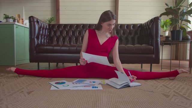 Multitasking freelance slim fit woman in sports jumpsuit sitting on side twine, analyzing financial data, making notes in organizer while doing stretching exercises and working on new project indoors.