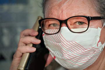 Elderly senior woman with glasses wearing hand made cotton mouth nose virus face mask, talking over her phone. Coronavirus covid19 outbreak prevention concept