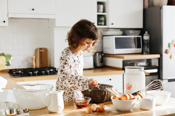Cute 5 years girl wearing dress preparing cake at the white modern kitchen.