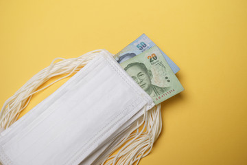 A pile of face mask with Thai Baht money isolated over yellow background. Concept during Corona virus outbreak in Thailand, shortage of face mask increase the demand in higher price.