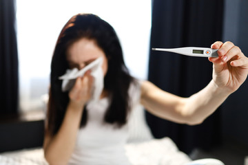 A young woman has a cold and high temperature while checking her body temperature with a digital thermometer.