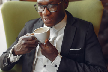 Black man in a cafe. Guy in a glasses. Man in a black suit. Businessman drinking a coffee.
