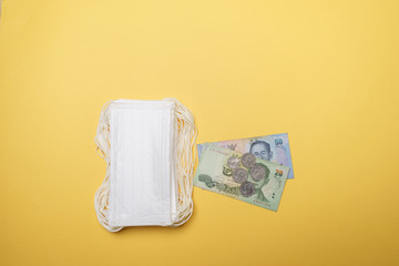 A pile of face mask with Thai Baht money isolated over yellow background. Concept during Corona virus outbreak in Thailand, shortage of face mask increase the demand in higher price.