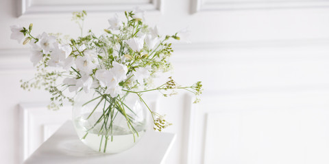 Bouquet of gentle bells in vase. Morning light in the room. Soft home decor, glass vase with white flowers on white wall background and on wooden table. Interior. Greeting card. Copy space. Banner