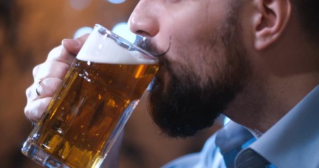 Extreme close-up of a handsome bearded man with a long twisted mustache, drinking beer with pleasure from large glass mug in dark pub. With free space for writing or info graphics. 4k slow motion.
