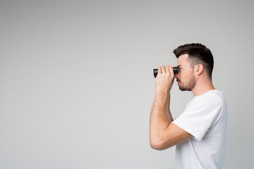 Young man isolated over background. Side view of guy looking forward through binoculars. Investigation or spy methods. Alone in room.
