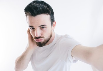 Casual man taking selfie in studio alone. Look straight and pose. Wear white shirt. Bearded man isolated over white background.