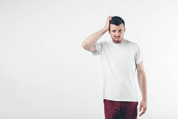 Young brunette man with beard dressed in white T-shirt. Guy сonfused and upset. He scratches his head with his right hand. Isolated over light background.