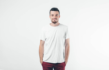 Young brunette man with beard dressed in white T-shirt. Guy confident and smiling. Hands holding in pockets of burgundy trousers. Isolated over light background.