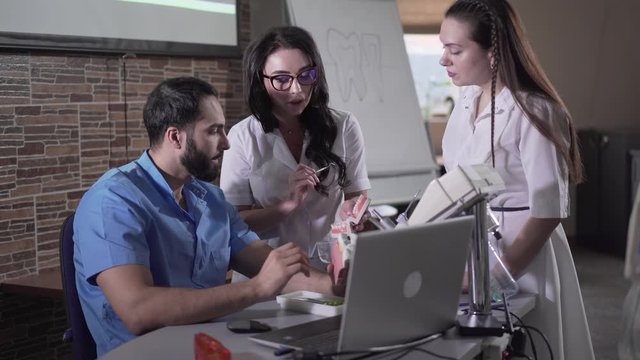 Modern Middle Eastern teacher explaining dentistry to students using laptop. Concentrated Caucasian women talking with smart stomatologist. Lifestyle, profession, odontology, medicine.