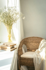 Cozy reading nook by the window with rattan wicker chair, delicate white gypsophila flowers, opened book and window light and shadows. Modern comfortable living room space. Slow living concept. - obrazy, fototapety, plakaty