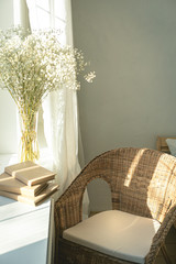 Cozy reading nook by the window with rattan wicker chair, delicate white gypsophila flowers, opened book and window light and shadows.Modern comfortable living room space - obrazy, fototapety, plakaty