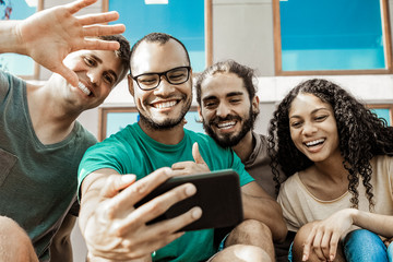 Cheerful friends during video chat via smartphone. Low angle view of happy multiethnic friends sitting together and talking during video chat outdoors. Communication concept