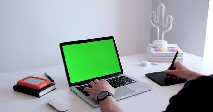 Medium close up of a man working, drawing, sketching on a laptop with a graphic tablet. Computer mockup with green screen chroma key display on a minimalist white work desk at the office with gadgets