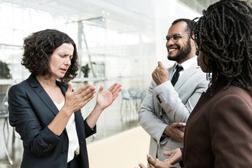 Excited multiethnic colleagues chatting outside. Business man and women standing at outdoor glass wall, talking to each other, gesturing, laughing. Business communication concept