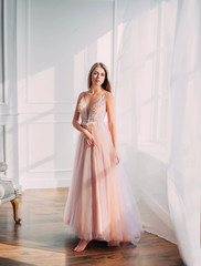 Beautiful young woman. Blonde long flowing hair. elegant airy peach beige ball gown. evening long pink trendy tulle dress. backdrop white window vintage classic room. Prom holiday image. Stylish look © kharchenkoirina