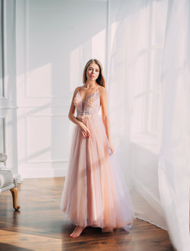 Beautiful young woman. Blonde long flowing hair. elegant airy peach beige ball gown. evening long pink trendy tulle dress. backdrop white window vintage classic room. Prom holiday image. Stylish look