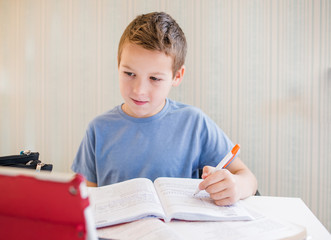 Naklejka premium 10-year-old boy schoolboy does distance learning lessons on a tablet while sitting at a table in a mask, the topic of distance learning of students in quarantine
