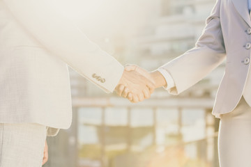 Business colleagues greeting each other in early morning. Business man and woman shaking hands in sunlight. City buildings in background. Agreement concept