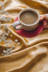 Сup of coffee in hands on a golden satin fabric with gypsophila flowers. Spring morning