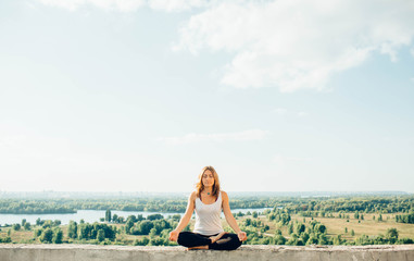 Naklejka na ściany i meble Young woman practices yoga outside. Calm peaceful girl sitting on parapet in lotus position. Her hands lowered to knees. Trees river and sky on background.