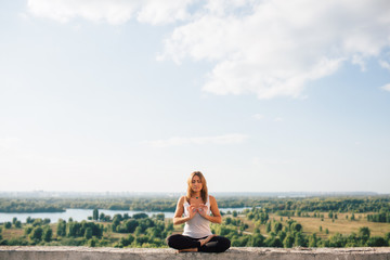 Naklejka na ściany i meble Young woman practices yoga outside. Girl calm and peaceful sits on parapet in lotus position. Her fingers fold into sign of infinity. Nature trees river and sky on background.