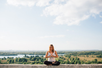 Naklejka na ściany i meble Young woman practices yoga outside. Calm peaceful girl sitting on parapet in lotus position. Her hands at chest level touch each other. Trees river and sky on background.