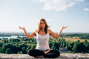 Young woman practices yoga outside. Calm blonde girl sitting on parapet in lotus position with...