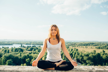 Naklejka na ściany i meble Young woman practices yoga outside. Calm smiling girl sitting on parapet in lotus position with closed eyes. Her hands lowered to knees. Trees river and sky on background.