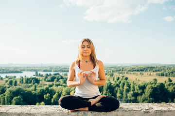 Fototapeta na wymiar Young woman practices yoga outside. Calm smiling girl sits on parapet in lotus position with close eyes. Her fingers fold into sign of infinity. Nature trees river and sky on background.