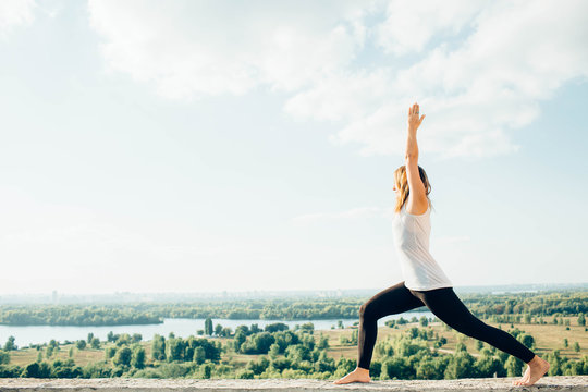Young woman practices yoga outside. Blonde girl standing in Warrior I posture (Virabhadrasana 1), full length on parapet on beautiful view. Trees river and sky on background.