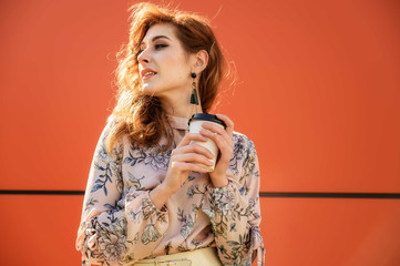 Photo in bright weather of a fashionable glamorous beautiful woman with long hair on an orange background in spring in the sun. Model stands outdoors