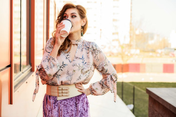 Photo in bright weather of a fashionable glamorous beautiful woman with long hair against the background of the spring city in the sun. Model is standing outdoors with a cup of coffee in her hands.