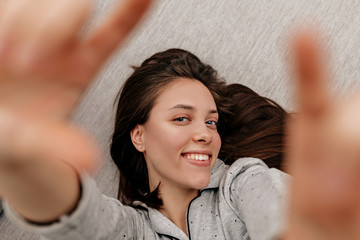 Happy lovable girl with dark hair wearing pajamas lying on the coach and making a selfie at home in...