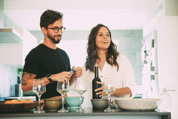 Happy cheerful couple waiting friend for dinner and cooking in kitchen. Young man and woman in...