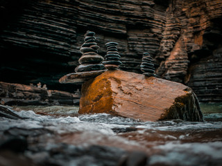 Piles of pebble stones balanced on top of a big rock. Shallow sea waves in foreground. Coastal image. Lots of textured and shiny rocks. 