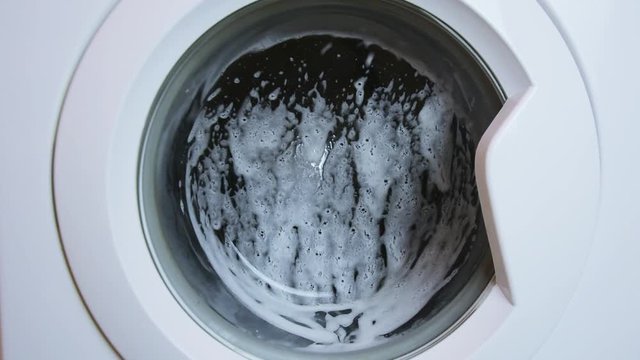 Washing clothing in generic domestic washing machine.Close up video of spinning washing machine drum with foam spraying door. 