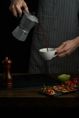Chef man hands holds coffee pot and pouring coffee into a white cup. Dark style photo.