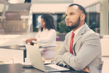 Pensive businessman with laptop thinking over startup strategy over cup of coffee. Young African American woman drinking coffee in background. Working in cafe concept
