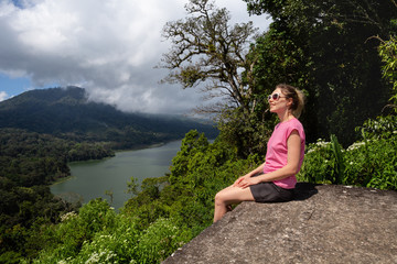 Fototapeta na wymiar Young traveling blonde woman is sitting on hill enjoying beautiful view of Tamblingan lake and mountains on Bali, Indonesia.