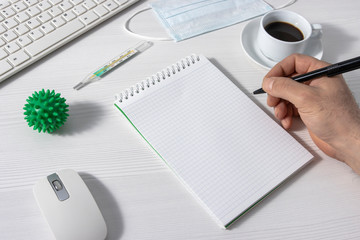 The man writes in an empty notebook. A coronavirus molecule, keyboard, mouse, thermometer, coffee, and medical mask on a white office Desk.