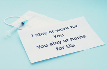 A piece of paper with printed text, asking doctors: "I stay at work for you, you stay at home for us", a medical mask and a syringe next to it. Selective focus. Covid19 coronavirus quarantine
