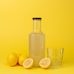 Bottle with lemon juice and a glass empty glass next to it and whole and sliced lemons isolated on a yellow background