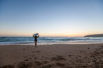 Fototapeta na wymiar Female surfer standing on beach at sunrise holding a surfboard
