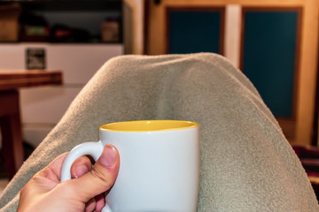 A first person perspective / point of view shot of a young Caucasian woman's hand holding a cup of hot beverage with her legs under a gray blanket in the living room