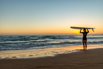 Fototapeta na wymiar Female surfer standing on beach at sunrise holding a surfboard