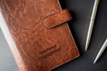 Rustic leathered Employee handbook on office desk with engraved text next to two steel pens on...