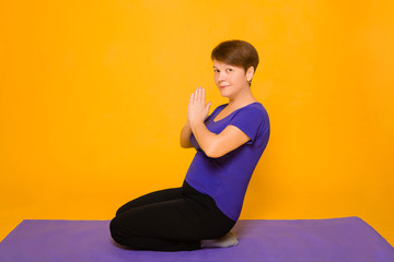 Fototapeta premium Aged woman doing yoga on a purple rug. Studio photo on a yellow background.