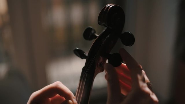 Young woman fiddler changing violin strings, tighten the string with the tuning peg at pegbox, moody low light scene at home
